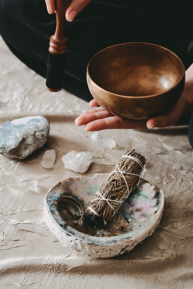 A Tibetan singing bowl in a person's hands and sage and crystals on the floor